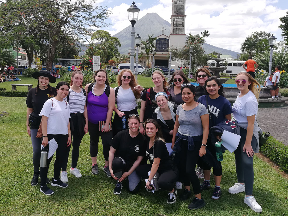 A group of students together in a tropical area with a small church in the background and a volcano in the distance