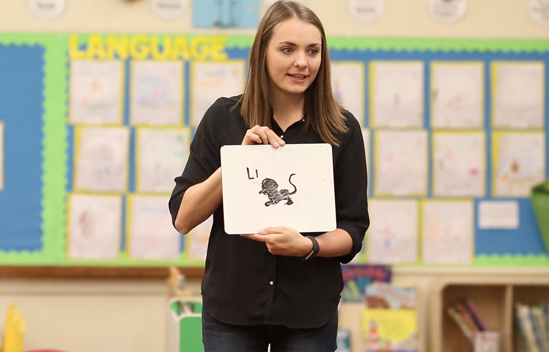 A teacher holding a small white board with an illustration of a lion asking for the proper spelling